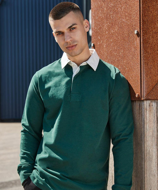 A man with a short haircut leans against a rust-colored panel, wearing a green long-sleeve shirt with a white collar from Front Row's FR104 collection. The background is a mix of blue and rust-colored surfaces.