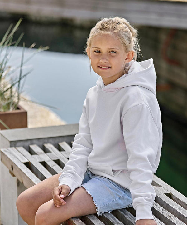 A young girl with blonde hair tied back sits on a wooden bench by the water, wearing a white Tee Jays Junior UNLABELLED hooded sweatshirt (5152B) and light blue denim shorts, looking at the camera with a neutral expression.