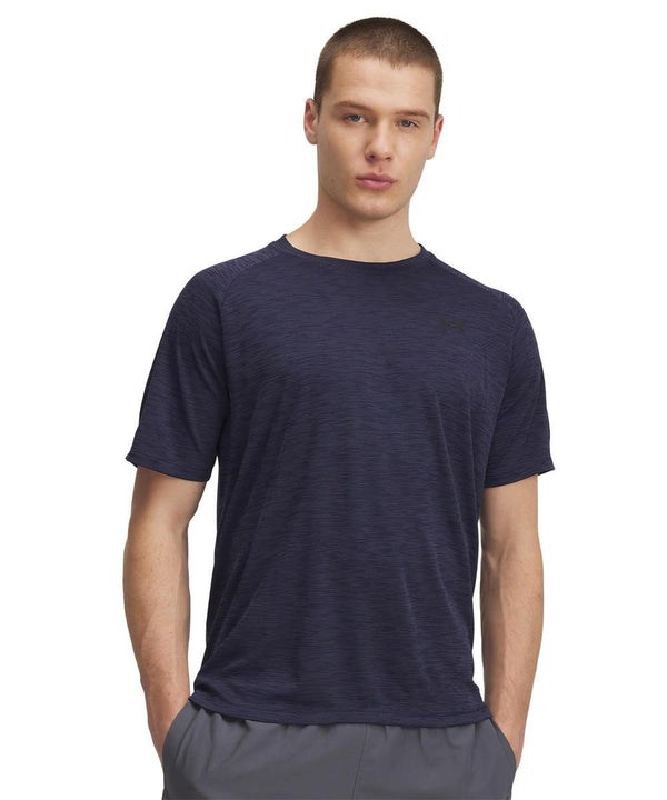 A young man with short hair stands with both hands in his pockets, wearing an Under Armour UA Tech textured T-shirt in dark blue and grey shorts against a plain white background.