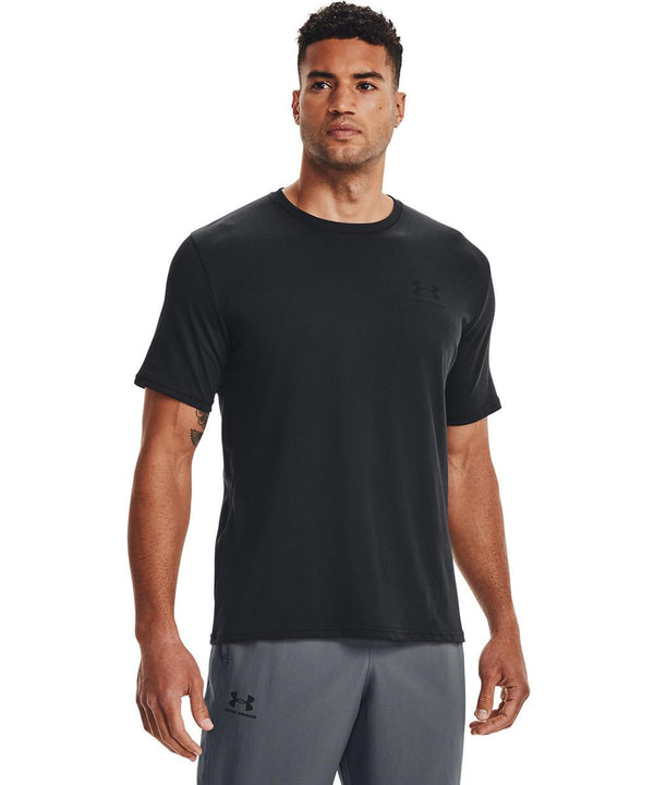 A man faces forwards wearing a black Under Armour UA Sportstyle short-sleeve T-shirt and grey athletic shorts against a plain white background.
