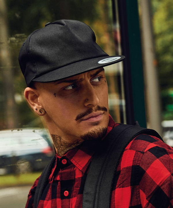 A man with tattoos and a goatee, wearing a black Flexfit By Yupoong YP047 cap and a red plaid shirt, looks to his left. He stands in front of a reflective window with blurred outdoor scenery.