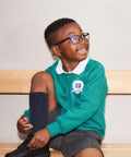 A young boy with glasses sits on a wooden bench, smiling and looking to the side. He is wearing a green school sweater with a Russell Europe logo, a white collared shirt, gray shorts, and dark knee-high socks. He appears to be adjusting his black 7620B.