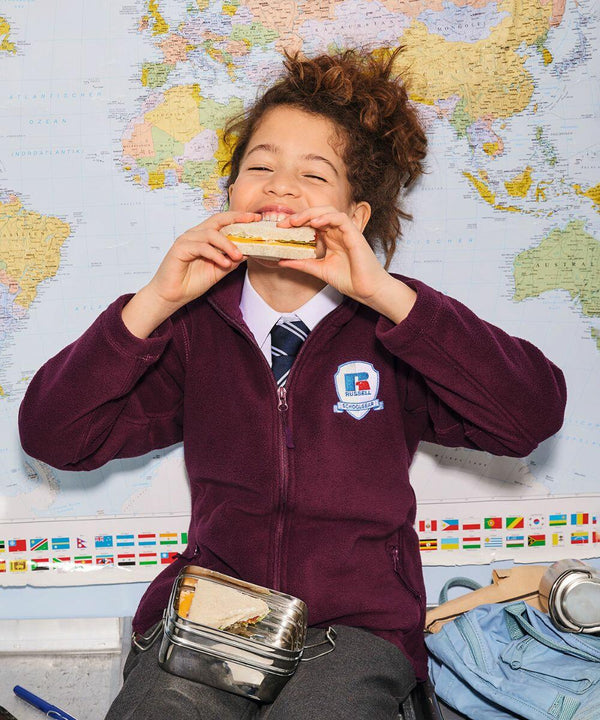 A cheerful child with curly hair and a school uniform eats a sandwich. They are sitting in front of a world map, holding a Russell Europe 8700B. Various small items and a pencil case are spread out on the table below the map. A badge on their sweatshirt reads "Student Ambassador.