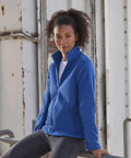 A person with curly hair is leaning against a railing while wearing a blue zipped-up Russell Europe 8700F jacket over a white shirt and jeans. The industrial background features a weathered metal structure. The person looks directly at the camera with a serious expression.