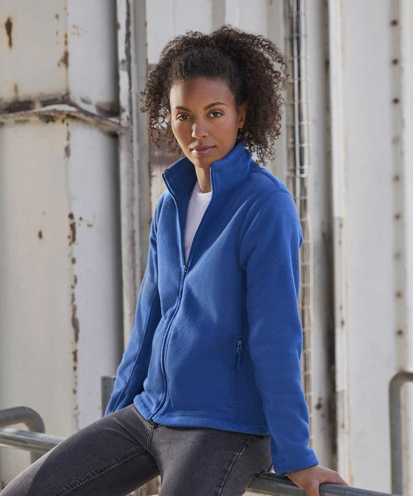 A person with curly hair is leaning against a railing while wearing a blue zipped-up Russell Europe 8700F jacket over a white shirt and jeans. The industrial background features a weathered metal structure. The person looks directly at the camera with a serious expression.