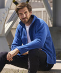 A man with short brown hair and a beard is sitting outdoors on a concrete step, wearing a bright blue Russell Europe 8700M zip-up jacket and black pants. The background features a metal structure and blurred urban elements. He appears relaxed, resting his hands on his knees.