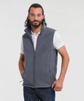 A man with long hair and a beard is wearing a gray Russell Europe 8720M over a white polo shirt and blue jeans. He stands with one hand in his pocket and the other resting on his hip, looking confidently at the camera against a plain background.
