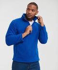 A man wearing a bright blue 8740M fleece jacket by Russell Europe and blue jeans stands against a plain background. He is pulling the zipper of the jacket up towards his chin, with a neutral expression. His head is slightly turned to his left.