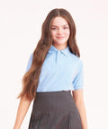 A young girl with long, wavy brown hair smiles at the camera. She is wearing an AWDis Academy AC004 light blue polo shirt and a dark gray pleated skirt. The background is plain and light-colored.