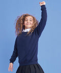 A young girl with curly hair stands against a blue background. She is wearing an AC01J navy-blue sweater by AWDis Academy and a pleated skirt. The girl smiles and raises one arm confidently towards the sky.