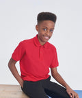 A young boy with a flat-top hairstyle is sitting and smiling. He is wearing a bright red AC04J polo shirt from AWDis Academy and black pants, and is seated on a light-colored wooden surface with a light blue and white background.