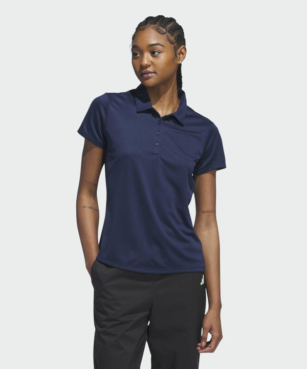 A person with braided hair is wearing an adidas AD003 navy-blue polo shirt and black pants, standing with one hand in their pocket, looking slightly to the side.
