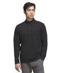 A man stands against a white background wearing the adidas Ultimate 365 classic quarter-zip in black and grey trousers, with his left hand in his pocket, looking slightly to the right.