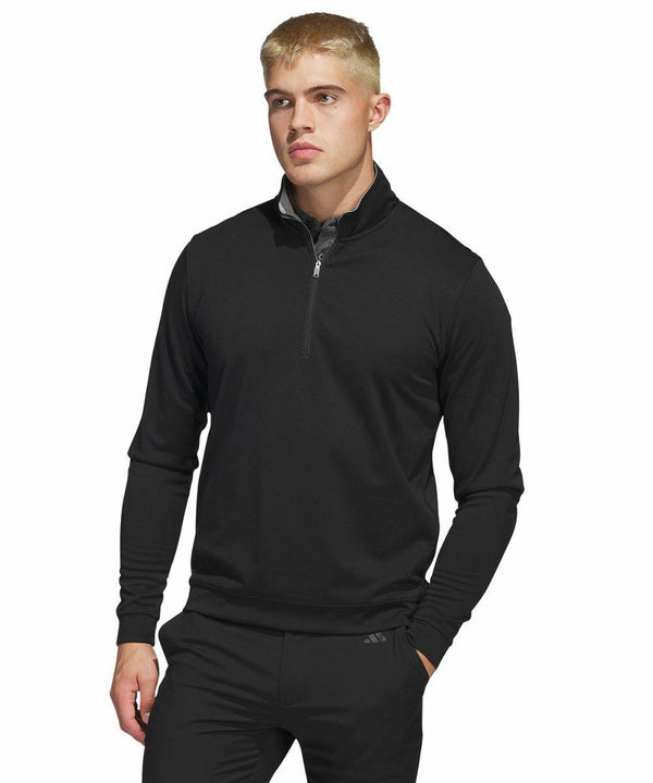 A man with short blond hair wears a black, long-sleeve, quarter-zip AD118 pullover from adidas and black pants. He stands against a white background, with his left hand resting in his pocket and a neutral expression on his face.
