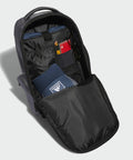 Image of an open black backpack displayed against a plain background. Inside, there's a pair of blue AD192 adidas socks, a pair of sunglasses tucked into a pocket, a zippered compartment, and a notebook or passport with a red cover. The interior is organized neatly.