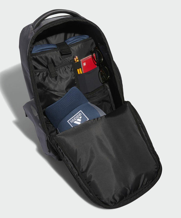 Image of an open black backpack displayed against a plain background. Inside, there's a pair of blue AD192 adidas socks, a pair of sunglasses tucked into a pocket, a zippered compartment, and a notebook or passport with a red cover. The interior is organized neatly.