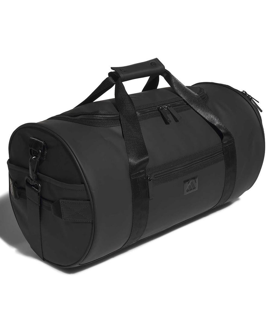 The adidas 1st tee duffle (26L) is a black cylindrical bag with two handles, a shoulder strap, and several zippered compartments, displayed on a white background.