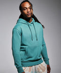 A person wearing a teal Anthem AM001 hoodie with the hood down and patterned pants stands against a plain background.
