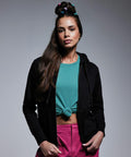 A young woman with long brown hair, styled with colorful hair ties, stands against a gray background. She wears an Anthem AM004 black hoodie over a tied teal shirt and pink pants, with her hands resting at her sides. She gazes confidently at the camera.
