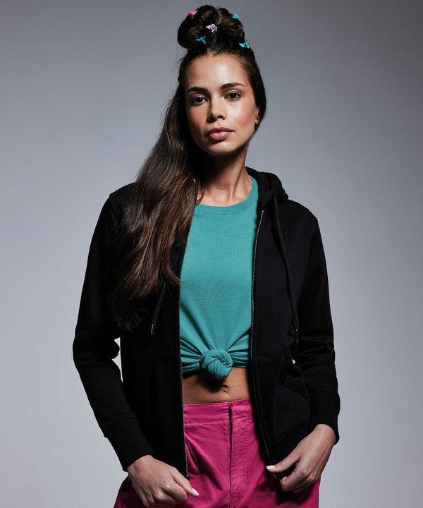 A young woman with long brown hair, styled with colorful hair ties, stands against a gray background. She wears an Anthem AM004 black hoodie over a tied teal shirt and pink pants, with her hands resting at her sides. She gazes confidently at the camera.