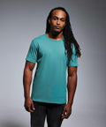 A person stands facing the camera, wearing a teal Anthem AM010 T-shirt and dark pants, with long braided hair.