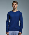 A young man stands facing forward against a plain grey background. He wears a long-sleeve, dark blue Anthem AM011 shirt and light grey pants. He has short, styled hair and a neutral facial expression.