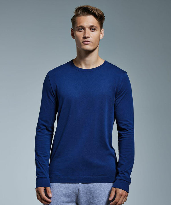 A young man stands facing forward against a plain grey background. He wears a long-sleeve, dark blue Anthem AM011 shirt and light grey pants. He has short, styled hair and a neutral facial expression.