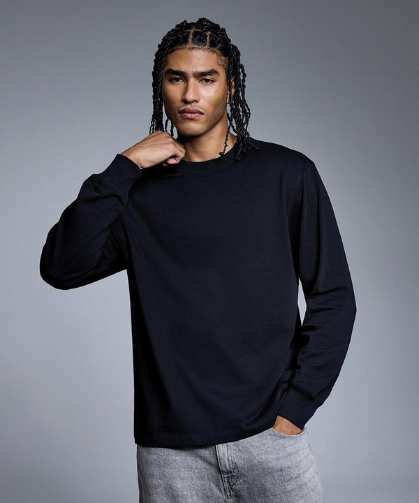 A person with long braided hair is wearing the Anthem Headline Heavy Long Sleeve T-Shirt in black, paired with light grey jeans, standing against a gradient grey background with one hand touching their neck.