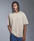 A person with long braided hair stands against a grey background, wearing the Anthem Amplify heavyweight t-shirt in beige by Anthem and blue jeans, with one hand in their pocket and a neutral expression.