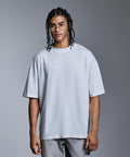 A person with plaited hair and glasses stands against a plain grey background, wearing the Anthem distortion washed T-shirt by Anthem and light grey trousers.