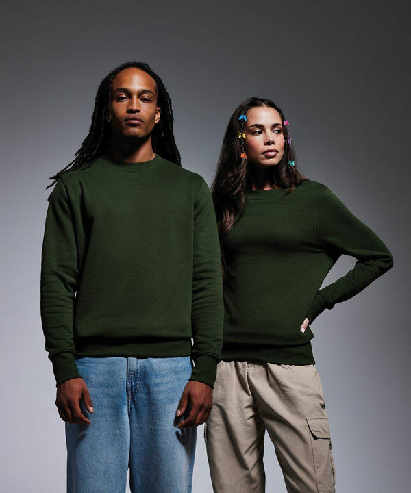 Two models stand side-by-side against a gray background, both wearing dark green sweatshirts from Anthem's AM020 collection. The person on the left has long dreadlocks and blue jeans, while the person on the right has long, wavy hair adorned with colorful hair clips and is wearing beige cargo pants.