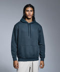 A young man with braided hair stands against a grey background, wearing the Anthem Studio hoodie by Anthem in dark teal and light-coloured trousers. He faces forward with a neutral expression.