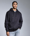 A person with plaited hair wears the Anthem Studio full-zip hoodie by Anthem over a grey shirt and light blue jeans, standing against a plain grey background with one hand in their pocket.