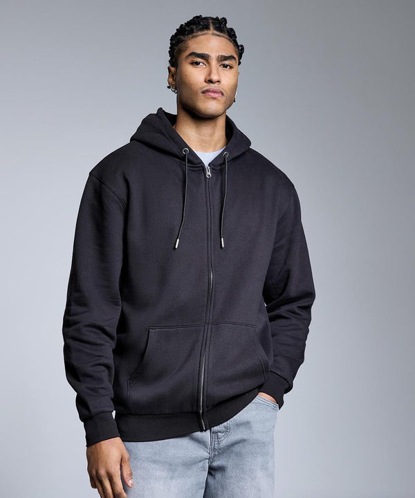 A person with plaited hair wears the Anthem Studio full-zip hoodie by Anthem over a grey shirt and light blue jeans, standing against a plain grey background with one hand in their pocket.