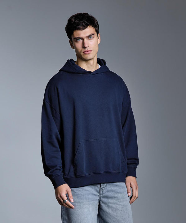 A young man stands against a plain grey background, wearing the Anthem heavy bass hoodie by Anthem with light blue jeans. He faces the camera with a neutral expression and one hand by his side.