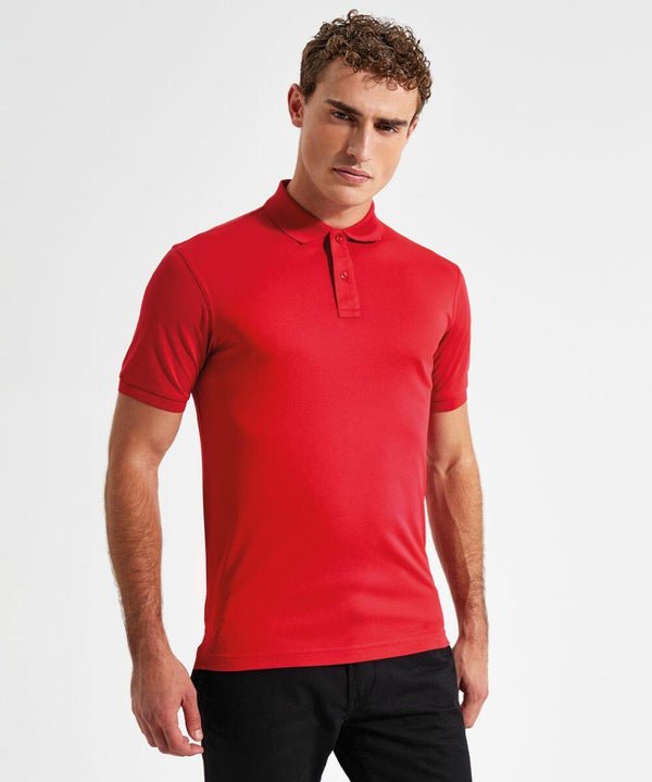 A man with short curly hair stands against a plain white background, wearing an AQ001 by Asquith & Fox bright red polo shirt and black pants. He has a serious expression on his face and his arms are relaxed by his sides.