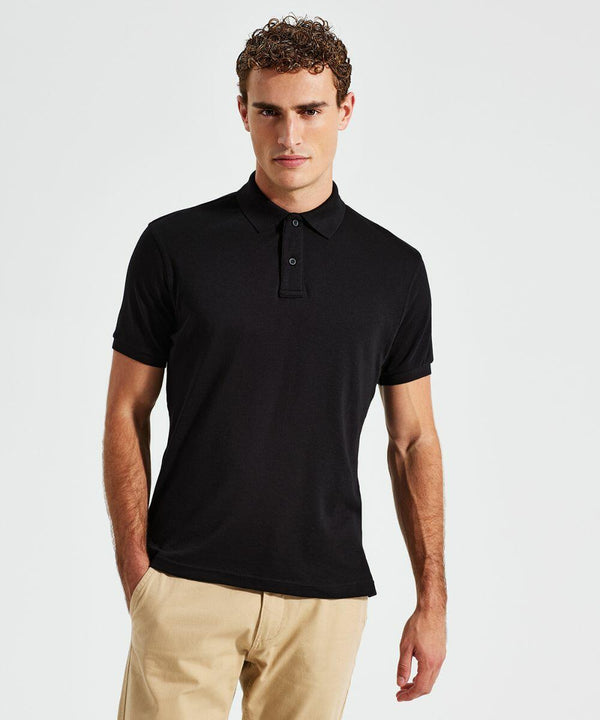 A man with short curly hair is wearing an Asquith & Fox AQ005 black polo shirt and beige pants. He has one hand in his pocket and stands against a plain white background.