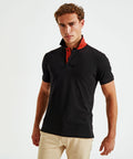 A man with curly hair wears an Asquith & Fox AQ012 black polo shirt with a red collar and beige pants, standing against a white background.