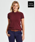 A woman smiles while wearing an Asquith & Fox AQ020 maroon short-sleeve polo shirt and dark blue pants with a brown belt. The image has a label indicating the shirt is made of 100% cotton.