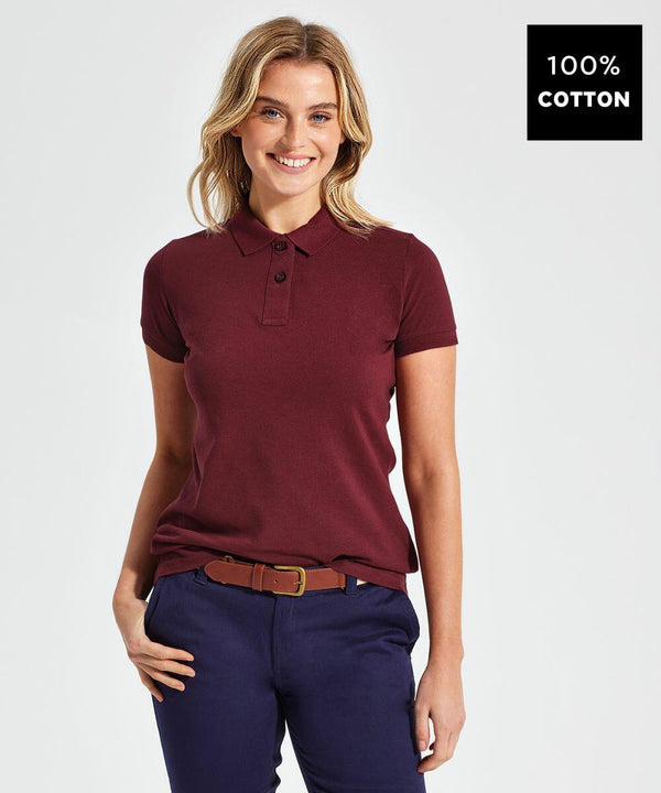 A woman smiles while wearing an Asquith & Fox AQ020 maroon short-sleeve polo shirt and dark blue pants with a brown belt. The image has a label indicating the shirt is made of 100% cotton.
