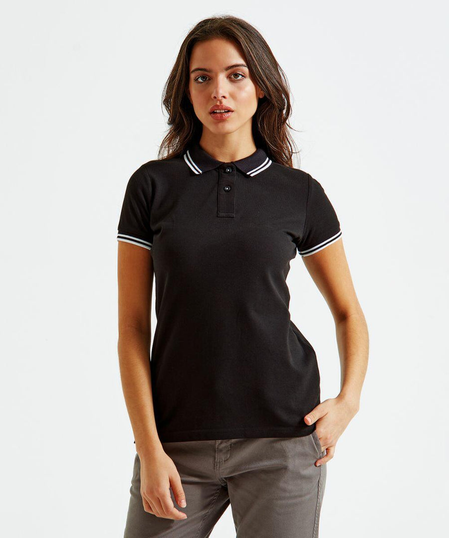 A woman with long dark hair, wearing an Asquith & Fox AQ021 black polo shirt with white trim on the collar and sleeves, and gray pants, stands against a white background. She looks directly at the camera with a neutral expression, with one hand resting on her hip.