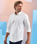 A man with light brown hair and a short beard is wearing an Asquith & Fox Men's cotton long-sleeve Oxford shirt with rolled-up sleeves, looking to his right against a light background featuring blue geometric shapes.