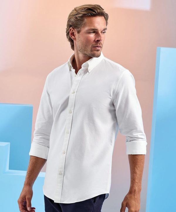 A man with light brown hair and a short beard is wearing an Asquith & Fox Men's cotton long-sleeve Oxford shirt with rolled-up sleeves, looking to his right against a light background featuring blue geometric shapes.