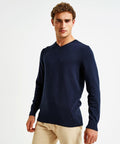 A person with short curly hair is wearing a dark blue Asquith & Fox AQ042 V-neck sweater and light beige pants. The individual is standing against a white background with a slight forward-facing posture and a neutral expression.