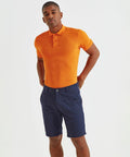 A man wears an orange Asquith & Fox AQ051 polo shirt and dark blue shorts, standing against a plain white background.