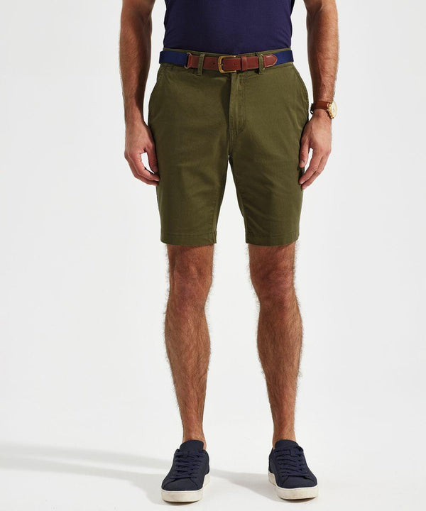 Man wearing an Asquith & Fox AQ058 navy blue polo shirt, green shorts with a brown belt, a wristwatch, and blue sneakers, standing against a white background.