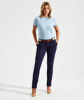 A person stands against a plain background, dressed in a light blue Asquith & Fox AQ060 polo shirt, navy blue pants, a brown belt, and beige shoes, with their hands in their pockets.