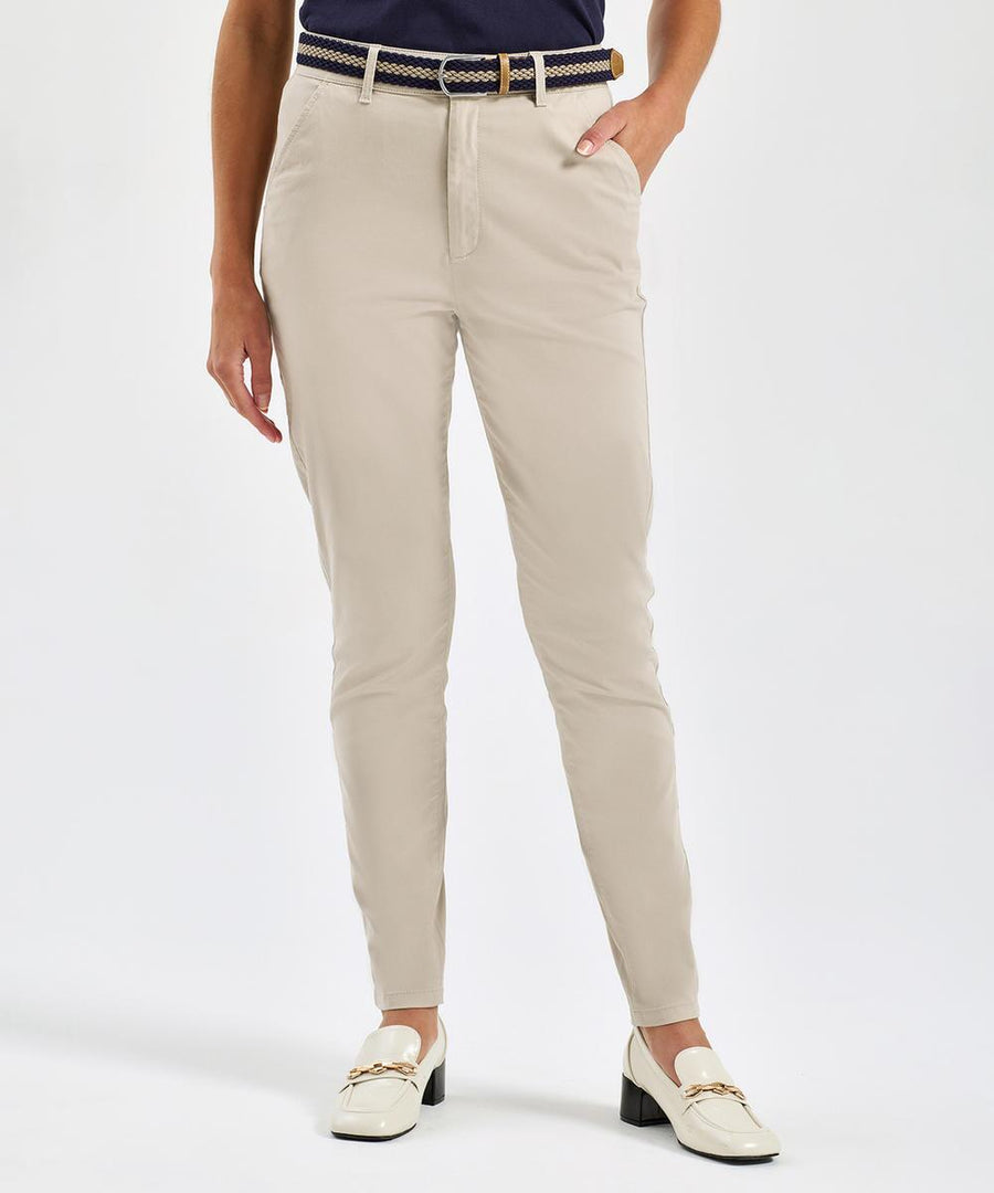 A woman models Asquith & Fox Women's Ultimate Chinos in light beige with a navy belt, dark top, and cream loafers. She stands against a white background, one hand in her pocket. The photo is cropped at the shoulders.