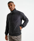 A person stands against a plain background, wearing an Asquith & Fox AQ200 black jacket with a zip-up front and their hand in one of the pockets. They have short hair and a neutral expression.