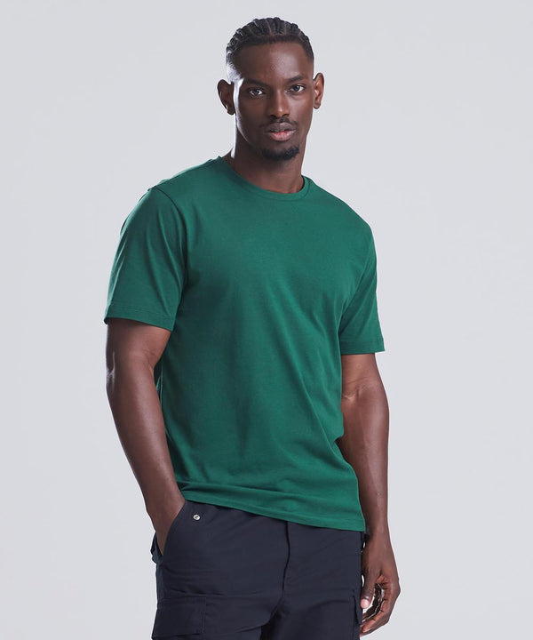 A man stands against a plain light background, wearing the AWDis 150 T green short-sleeve t-shirt by AWDis and black cargo trousers. He faces forward with one hand in his pocket and a neutral expression.